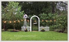 a white gate with an arch and windmill in the middle surrounded by green grass, trees, and string lights