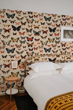 a bed with white sheets and butterflies on the wall behind it, next to a small side table