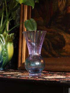 a purple glass vase sitting on top of a table next to green plants and paintings