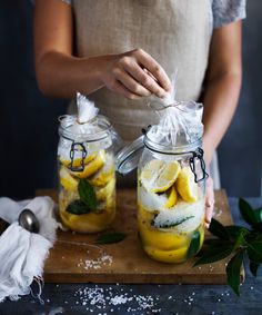 a woman holding two mason jars filled with lemon slices and mint leaves on top of a wooden cutting board