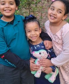 three children standing next to each other with one holding a stuffed animal and smiling at the camera