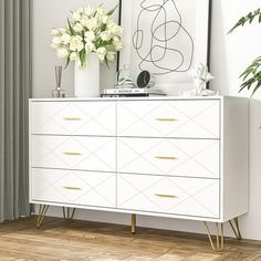 a white dresser with gold legs and flowers on top in a living room area next to a window