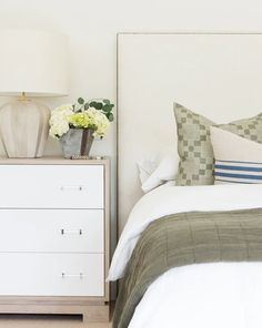 a bed with white sheets and pillows next to a nightstand with flowers on the night stand