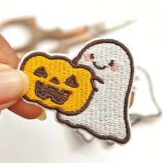 a hand holding a small embroidered patch with a ghost and pumpkin on it's face