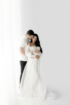 a bride and groom embracing in front of a white backdrop with the light coming from behind them