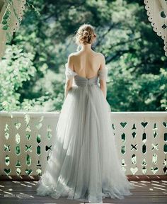 a woman in a white dress standing on a porch looking out at the trees and bushes