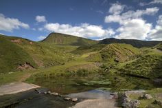 there is a small lake in the middle of this mountain range with green hills behind it