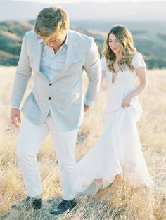 a young man and woman are walking through the grass in an open field, dressed in white