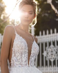 a woman in a white wedding dress standing next to a fence with the sun shining on her face