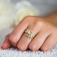 a woman's hand with a gold ring on top of her finger and white flowers in the background