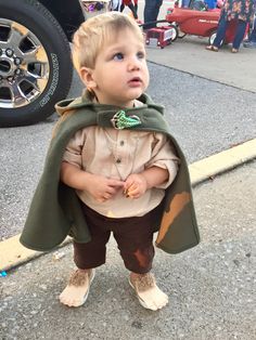 a little boy dressed in a green cape standing on the street with his hands behind his back