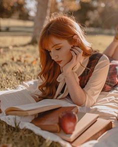 a woman laying in the grass reading a book and talking on her cell phone while holding an apple