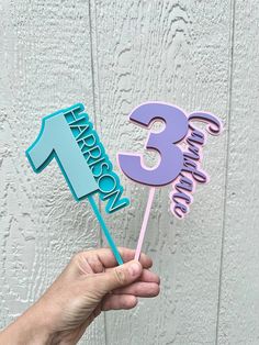 a hand holding a cake topper with the number three in blue, purple and pink