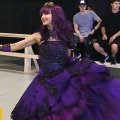 a woman in a purple and black dress dancing with other people sitting on the stage behind her