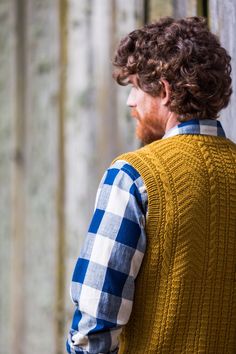 a man with curly hair wearing a yellow vest and plaid shirt standing in front of a wooden fence