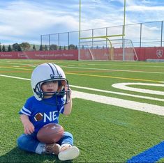 a little boy sitting on top of a football field wearing a helmet and holding a football