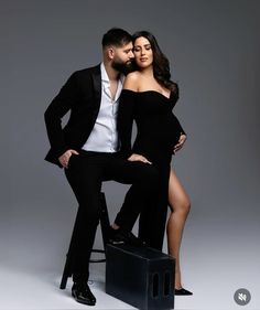 a man and woman posing for a photo in front of a gray background, with one leg on the chair