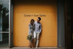 a man and woman standing next to each other in front of a yellow wall with writing on it