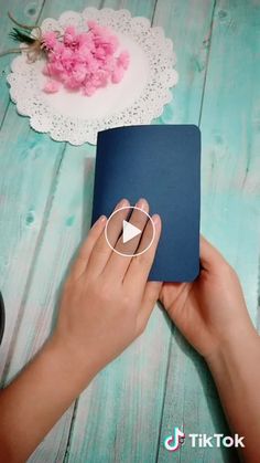 a person holding a book on top of a wooden table with pink flowers in the background