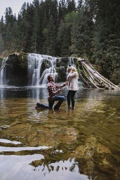 a man and woman holding hands while standing in the water near a waterfall with trees