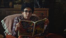 a young man sitting in a chair reading a comic book while wearing eyeglasses