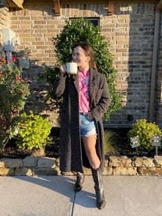 a woman standing on the sidewalk drinking from a mug while wearing short shorts and boots