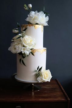 a three tiered cake with white and gold flowers on the top, sitting on a wooden table