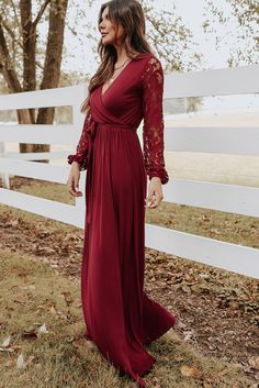 a woman standing in front of a white fence wearing a long red dress with lace sleeves