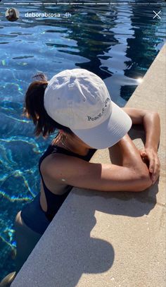 a woman sitting on the edge of a swimming pool wearing a white hat and black tank top