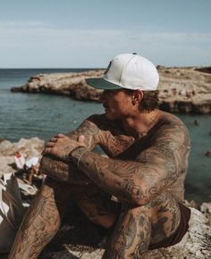 a man with tattoos sitting on rocks near the ocean