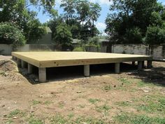 a wooden platform sitting in the middle of a yard next to trees and dirt ground