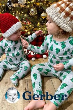 two babies sitting on the floor in pajamas and hats next to a christmas tree with presents