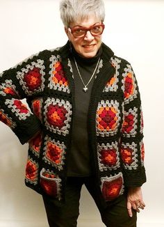 an older woman standing in front of a white wall wearing black pants and a colorful sweater