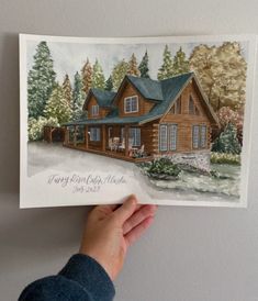 a person holding up a card with a drawing of a log cabin in front of it