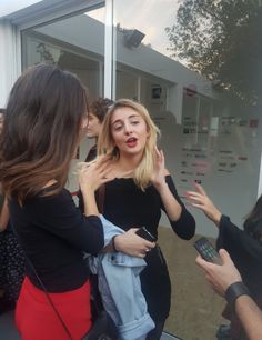 two women are talking to each other in front of a store window with people looking at them