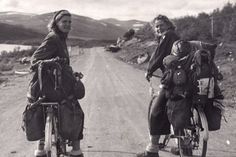 two women riding bikes down a dirt road with bags on the back and one woman standing next to her