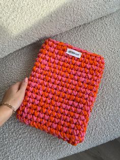 a hand holding an orange and pink crochet purse on the ground next to a couch