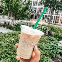 a person holding up a starbucks drink in front of some trees and bushes with buildings in the background