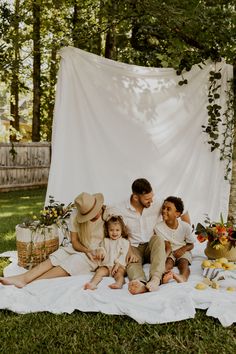 a family sitting on the grass in front of a white backdrop with flowers and plants