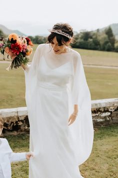 a woman in a white wedding dress walking with a flower crown on her head and veil over her shoulder