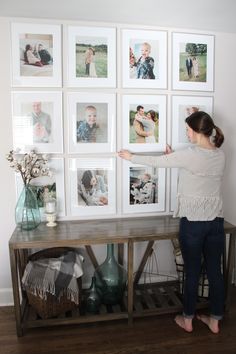 a woman standing in front of a table with pictures on the wall behind her and holding onto a shelf