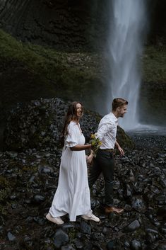 a man and woman are standing in front of a waterfall holding hands with each other