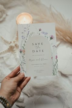 a person holding up a save the date card in front of some feathers and flowers