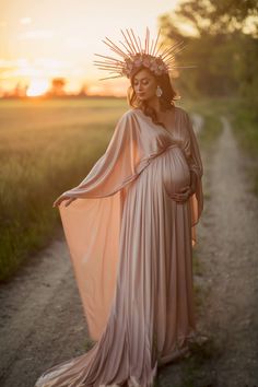 a pregnant woman wearing a dress and headpiece standing on a dirt road at sunset