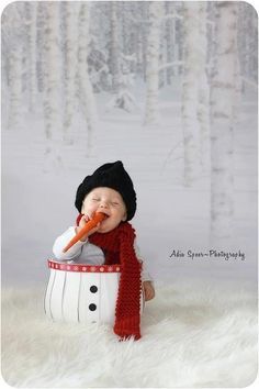 a baby wearing a hat and scarf eating a carrot while sitting in a snowman bucket