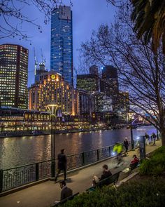 people are sitting on benches near the water in front of some tall buildings at night