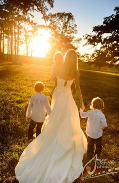 a woman in a wedding dress holding hands with two small children walking towards the sun