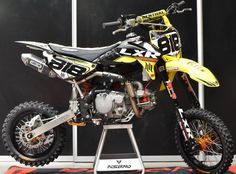 a yellow dirt bike on display in front of a black wall with red and white lettering