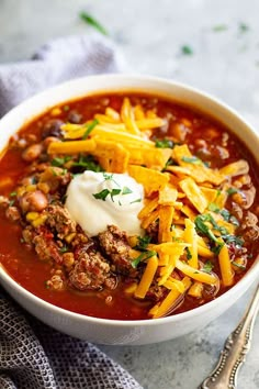 a bowl of chili with cheese and sour cream on top, next to a spoon