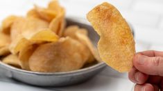 a hand holding up a piece of fried food in front of a metal bowl filled with potato chips